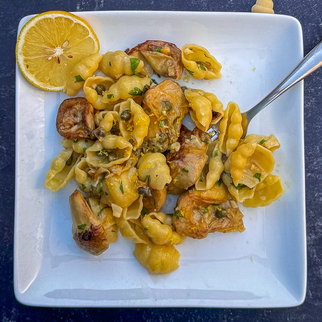Pasta dish with artichokes on a white plate with a lemon wedge, on a dark surface.