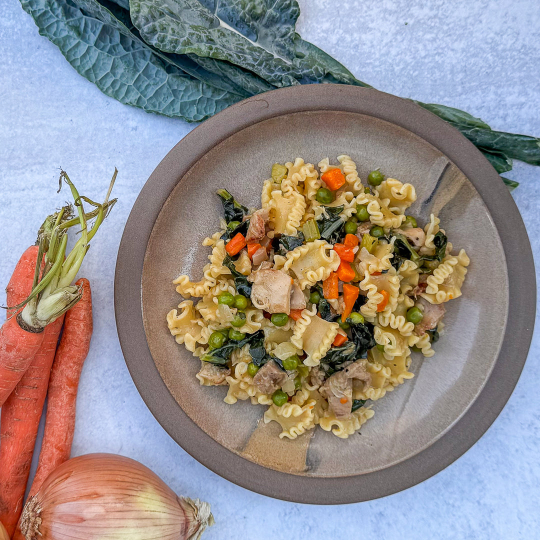 Pasta dish with vegetables on a plate, surrounded by carrots, onions, and kale.