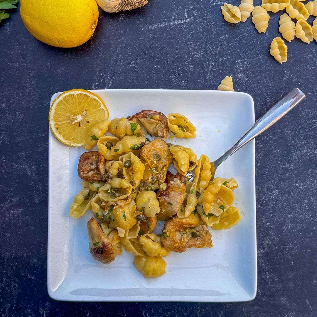 Pasta dish with artichokes on a white plate with a lemon wedge, on a dark surface.