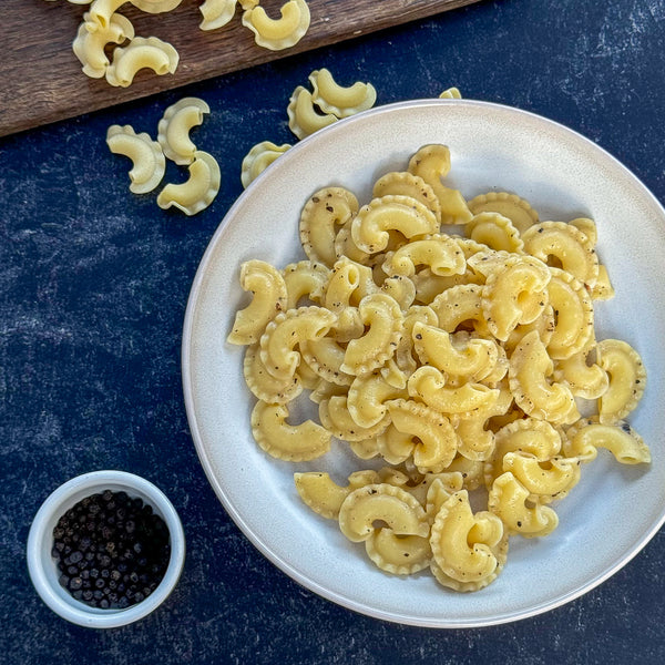 White plate with creste di gallo  pasta on a blue surface, small bowl of black pepper nearby.