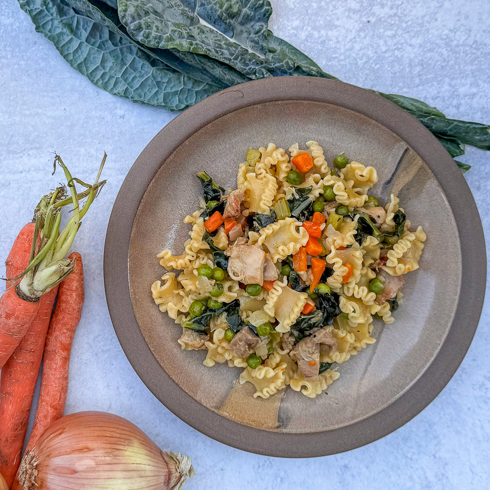 Pasta dish with vegetables on a plate, surrounded by carrots, onions, and kale.